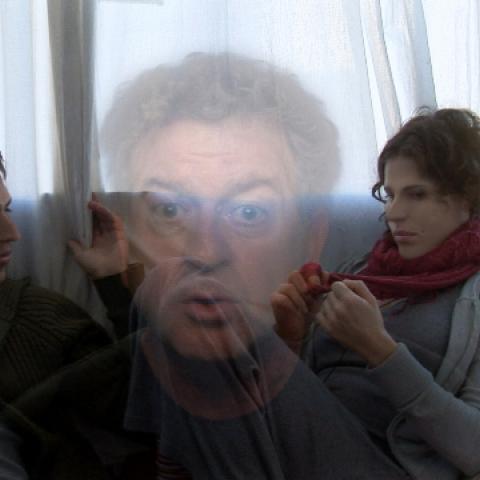 A man and a woman are sitting on the sofa talking. In the foreground, Avi Mograbi's face can be seen transparently in close-up.