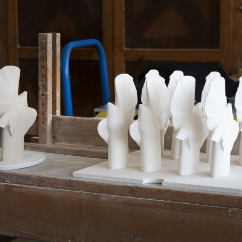 Auf einer Holzbank mit weißem Staub bedeckt stehen in Reihen ein Dutzend weißer Porzellantauben. Links von ihnen steht eine weitere, die gräulich und ein wenig rauher wirkt.