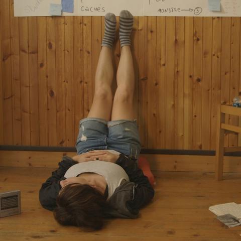 Filmstill: Eine Frau liegt in einem holzgetäfelten Raum auf dem Fußboden. Die Beine hat sie an der Wand hochgelegt. Sie trägt Socken und eine kurze Jeans-Shorts.