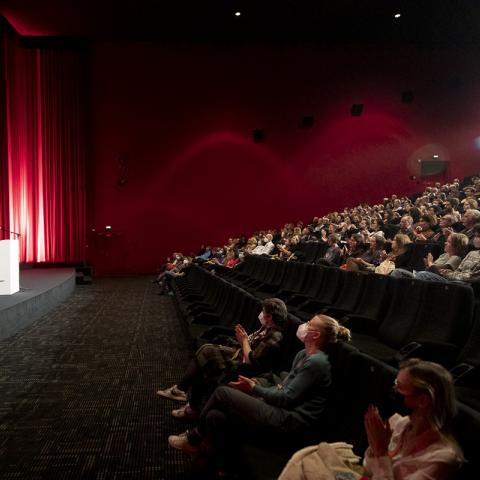 Cinema audience is listening to festival director Christoph Terhechte and moderator Julia Weigl who are talking to each other on stage in front of the screen.