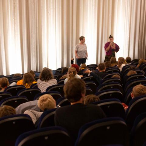 Kino bei einer Schulvorstellung von DOK Leipzig: Schüler*innen verschiedenen Alters hören der Regisseurin Susanne Kim zu, die vorne vor der Leinwand steht und in ein Mikrofon spricht.