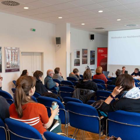 Ein Veranstaltungsraum, in dem verteilt mit Mindestabstand mehrere Personen sitzen und drei Vortragenden zuhören. Auf der Leinwand hinter den Vortragenden steht der Schriftzug: Methoden der Nachbereitung.
