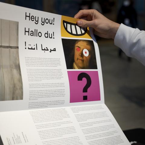Eine Hand hält ein großes aufgeklapptes Faltblatt, auf dem in der Mitte groß "Hallo Du! Hey You!" steht.