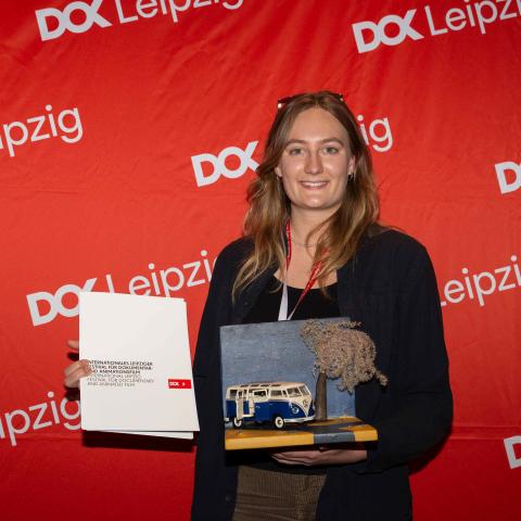 Filmemacherin Johanna Seggelke präsentiert vor einer Leinwand mit vielen DOK Leipzig Logos ihre Preisurkunde und Preisstatue. Die Statue besteht aus einer Platte, auf der ein VW-Bus neben einem Baum parkt.