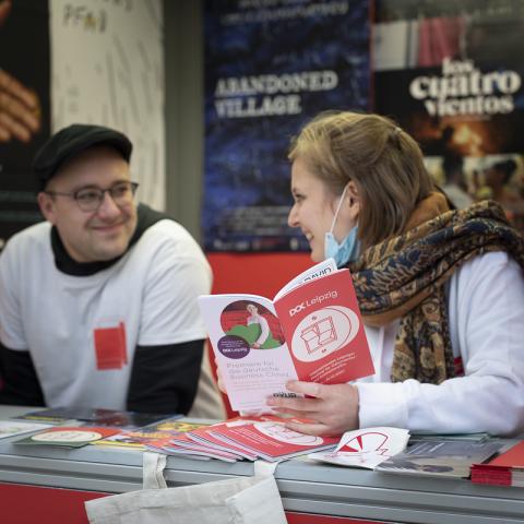 In der mit Postern geschmückten Dok box stehen zwei Personen mit Dok Pullovern und schauen gemeinsam ins Programmheft. Darin scheint etwas sehr lustig zu sein.