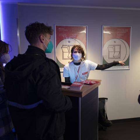 An einem Tisch voller DOK Leipzig Programme sitzt eine Frau mit DOK Leipzig T-shirt und weist den Personen vor dem Tisch den Weg