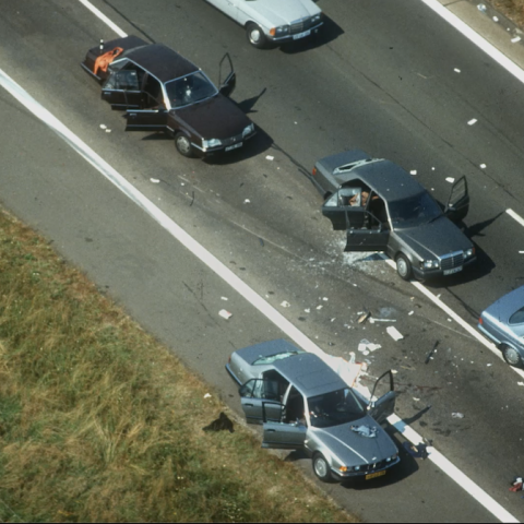 A bird’s eye view on a street where five cars stand in the middle of the street, all with doors open. The two cars in the centre have broken windows. Shards lie around.