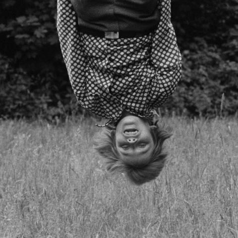 Ein schwarz-weiß Bild, über einem Weizenfeld hängt eine jugendliche Person in einem gepunkteten Hemd kopfüber in das Bild hinein. Die Haare stehen ganz zu Berge, sie lacht ausgelassen