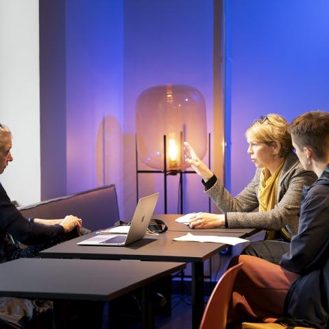 Three women sit together at a table in a productive atmosphere and talk. A laptop is on the table. The person in the middle gestures, the two people to her left and right look at her with interest.