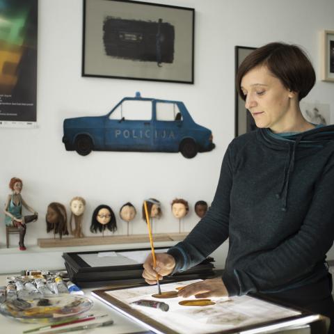 A woman with a short pageboy hairstyle stands at a work table in a bright room. She brushes with her right hand on a drawing board illuminated from below.