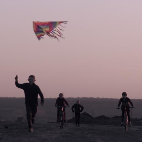 A wide area or street at dusk: four children move towards the camera. Two are running, two are riding their bicycles. Above them flies a kite, which the child on the far left holds by the string.