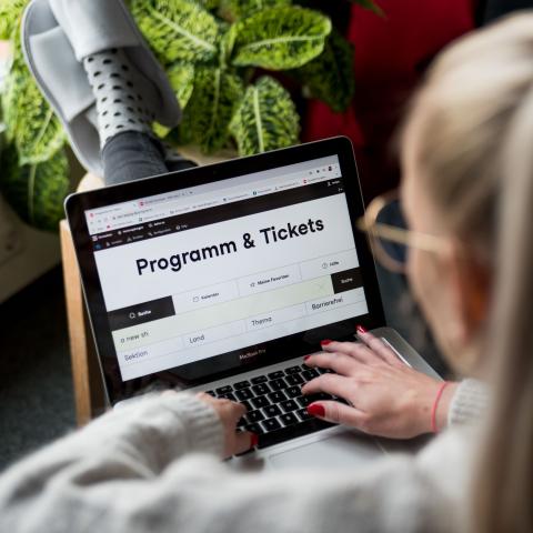 Eine Junge Frau in einem grauen Wollpullover ist von hinten zu sehen. Sie hat blondes Haar, rote Fingernägel und trägt eine Brille. Sie sitzt vor einem Laptop und tippt auf die Tastatur. Auf dem Bildschirm wird die Programm-Website von DOK Leipzig angezeigt. Die Füße in gemütlichen Hausschuhen hat sie bequem auf einem Hocker abgelegt. Im Hintergrund steht eine große Pflanze.