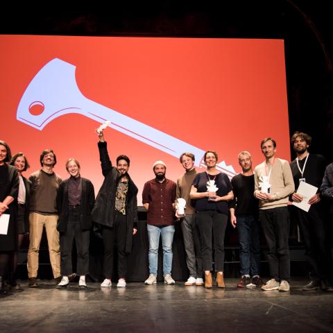 A group of 13 people is standing on a stage. All of them look towards the audience and smile. Some are holding small white porcelain dove figurines in their hands.
