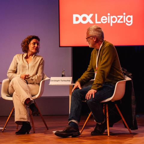 Eine Frau und ein Mann sitzen nebeneinander auf einem Podium. Sie haben sich einander zugewandt und sprechen mit einander. Im Hintergrund ist ein Bildschirm mit dem Logo von DOK Leipzig.