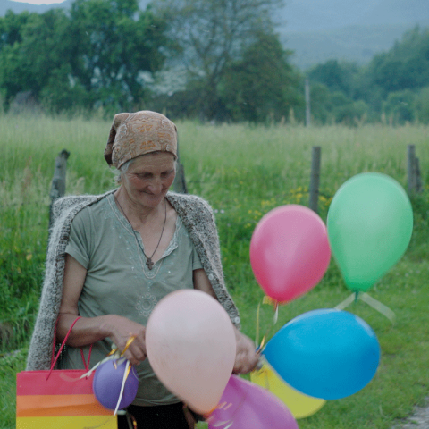 Eine ältere Frau steht vor einem Feld und hat ein Bündel an bunten Luftballons in der Hand. die um sie herumschweben.