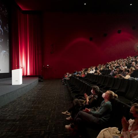 A crowded cinema hall. In front of the screen stand the director of DOK Leipzig Christoph Terhechte and moderator Julia Weigl.