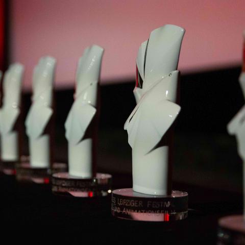 Eight white porcelain statuettes in the abstract shape of doves are lined up in front of a cinema screen. Some of the figurines have golden and some silver parts.