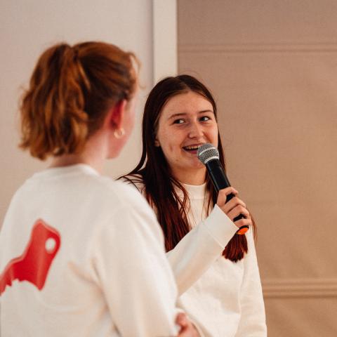 Eine Interviewsitutation: Zwei jugendliche Mädchen stehen nebeneinander und schauen sich an. Ein Mädchen spricht in ein Handmikrofon. Beide tragen Pullover mit dem Dok Leipzig Logo.