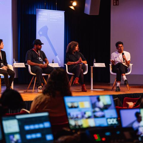 Four persons (two women, two men) are sitting on a panel. The man at the right is talking into a handheld microphone, the others are listening. In the front, the monitors of a live-editing suite light up in the dark.