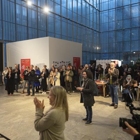 Many people stand in a large room with high glass fronts. They are gathered in a semicircle on front of a stage and clap.