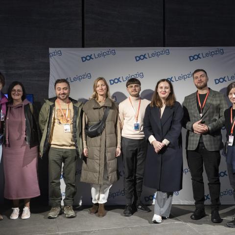 Eight persons (five women and three men) stand in front of a logo wall with the DOK Leipzig logo and pose for a group picture.