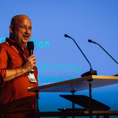 A men with a bald head and in an orange shirt stands at a speech desk and speaks into a microphone that he is holding in his hand.