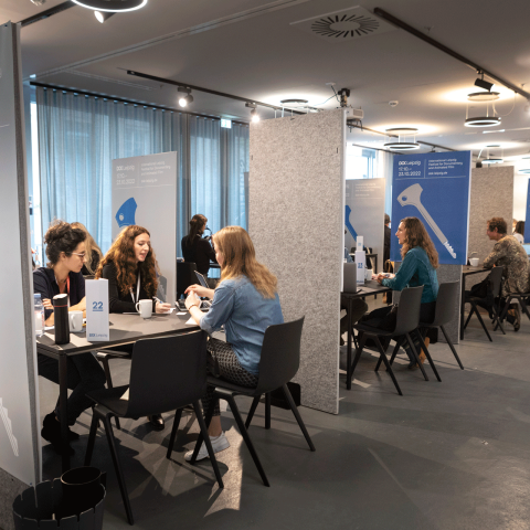 Eine Reihe von kleinen Konferenztischen, zwischen den Tischen stehen mobile Raumtrenner. An den Tischen sitzen sich jeweils 1 bis 2 Personen gegenüber und sind in Gespräch vertieft.