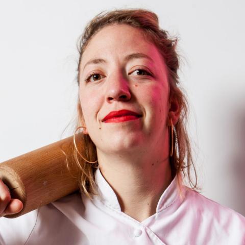 Portrait einer Frau, die einen Kochkittel trägt. Sie hat ein Nudelholz lässig auf ihre Schulter gelegt und schaut leicht lächelnd direkt in die Kamera.