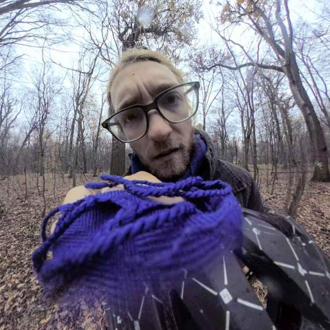 Ein junger Mann steht in einem Wald voller Herbstlaub und hat den Kopf zur Seite geneigt. Er blickt direkt in die Kamera.