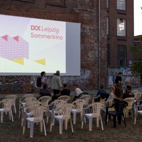 Mehrere Personen nehmen auf weißen Gartenstühlen Platz, die auf einer Wiese stehen. Sie schauen auf eine Leinwand, die an einer Backsteinwand befestigt ist. Es dämmert. Auf der Leinwand steht 'DOK Leipzig Sommerkino'.