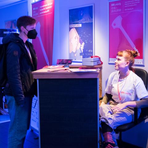 An einem Counter sitzt ein Volunteer, vor dem Counter steht eine Person in Jacke und mit Maske