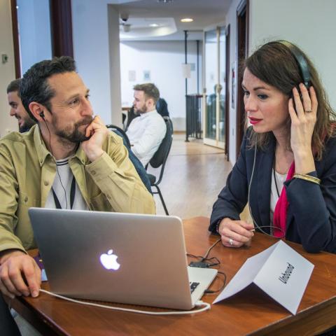 Two persons sit at a small table, the woman (at the right) wears earphones and looks at a laptop. The man (at the left) watches her.