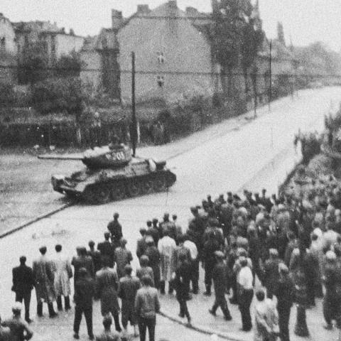 Schwarz-Weiß-Aufnahme: Eine Menschenmenge bewegt sich auf einen Panzer zu, der an einer Straßenecke steht.