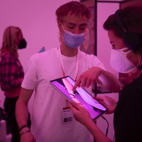 Ein Volunteer in der pink beleuchteten Neuland-Ausstellung zeigt einer Besucherin, wie das XR-Projekt auf einem iPad funktioniert