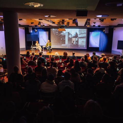 A large room for events. Two people are sitting on a podium. They are speaking in front of a large audience. Behind them is a screen showing clips from a film. 