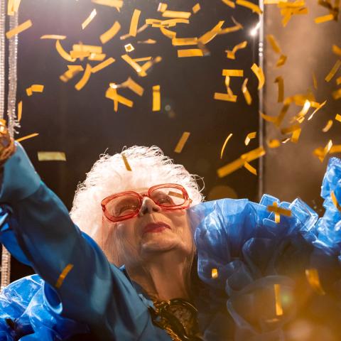 Older lady with oversized red glasses, blue costume and many frills throws cocky confetti in the air in front of glitter curtain