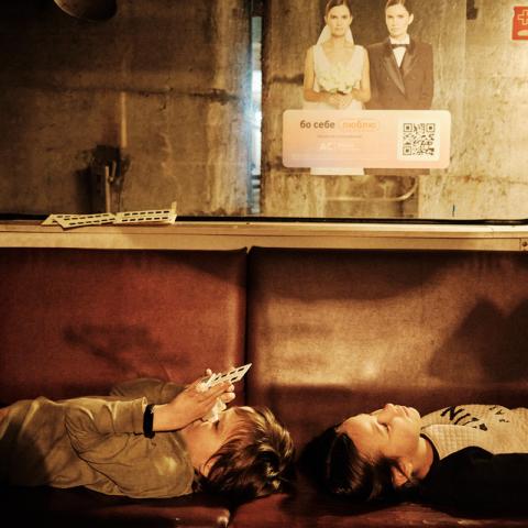Two children lying relaxed on a bench of a standing subway car. A view out of the carriage window reveals the wall of a subway tunnel.