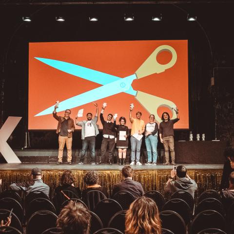 Sieben Personen stehen auf einer Bühne und halten Preisstatuetten in die Höhe. Im Hintergrund das Festivalmotiv von DOK Leipzig 2023: eine riesige Schere.