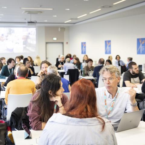 Ein Konferenzraum mit mehreren Tischen, die in Reihen hintereinander stehen. An jedem Tisch sitzen drei bis vier Personen, die miteinander im Gespräch sind.