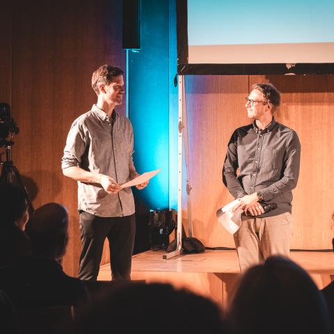Two men smile at each other in front of an audience in a wooden-paneled room. Both are holding certificates in their hands.