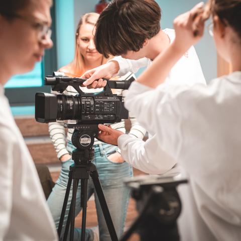 Eine Person setzt eine Kamera auf ein Stativ. Drei Personen schauen dabei zu.