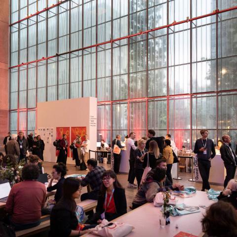 The festival centre, a large, bright room with a window front. Many people are busy sitting at tables or standing together, engaged in conversation.