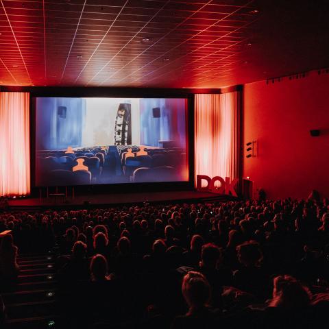 Ein voll besetzter Kinosaal, in atmosphärisches rotes Licht getaucht. Auf der Leinwand ist ein Ausschnitt aus dem Festivaltrailer zu sehen: kleine rote Gewichte, die ebenfalls in einem Kino sitzen.