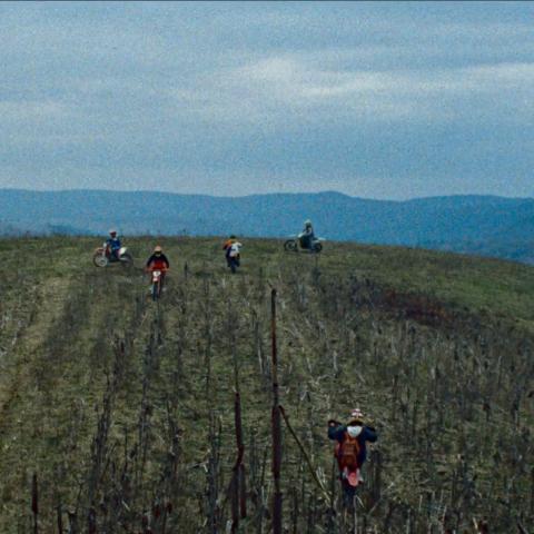 Wide shot of a grassy hill. Mountains are outlined in the background. Five people are driving up the hill on mopeds.