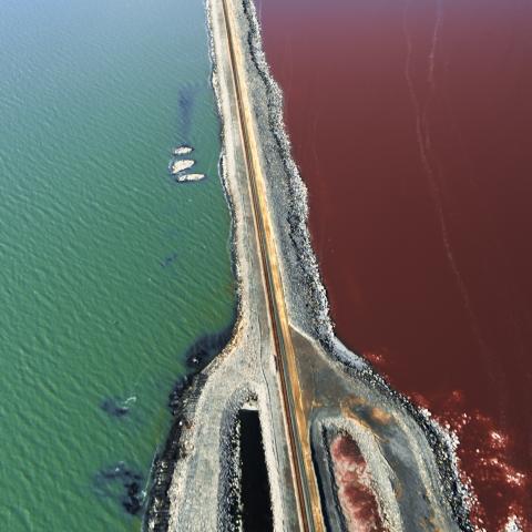 Aerial view of a road winding its way through a large stretch of water. The water is green-blue on one side of the road and rust-red on the other.