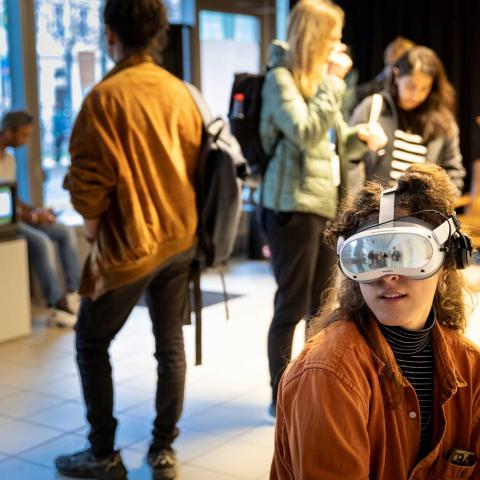 In einem Raum voller Menschen ist eine Person im Fokus, die eine verspiegelte VR-Brille trägt