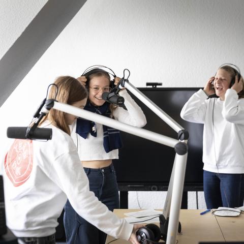 Drei Jugendliche in DOK Spotter Pullovern und mit Kopfhörern stehen um Radiomikrophone herum und lachen miteinander