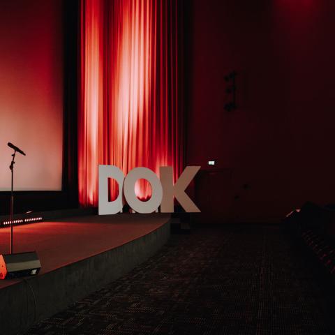 Eine Bühne vor einer Kinoleinwand. Darauf ein Mikrofonständer. Im Hintergrund ein roter Vorhang und große „DOK“-Buchstaben als Deko.