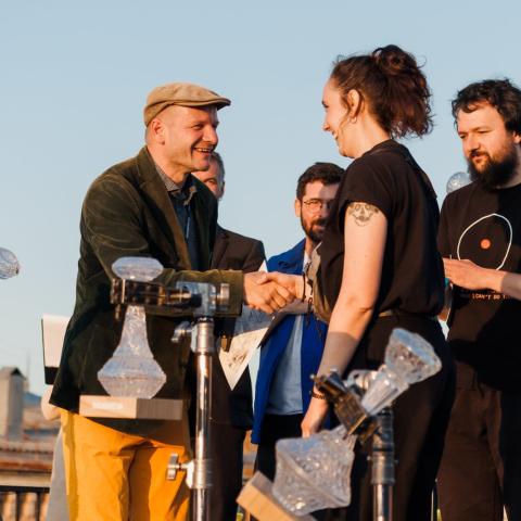 A small stage on a rooftop terrace. A young woman smiles and shakes the hand of a man who is presenting her with a certificate. Two other men stand in the background, also smiling.