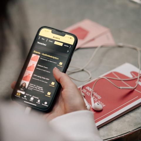 In Nahaufnahme ist eine Hand zu sehen, die ein Smartphone hält. Auf dem Display ist die App Greta geöffnet. Zu lesen ist: "Audiodeskriptionen". 